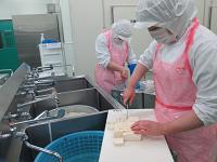 流しに渡したまな板の上で、ピンク色のエプロンをつけた作業者が豆腐を賽の目切りにしている写真