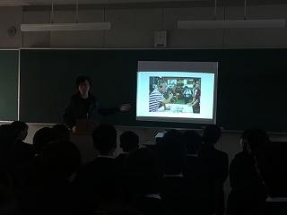 黒板に写真の載ったスライドを表示させて話している講師と、それを聞いている学生たちの写真