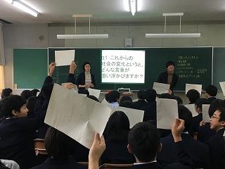 黒板の前に立つ二人の講師に向かって、白い紙を掲げている生徒たちの写真