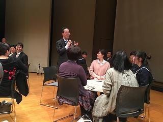 立って話をしている男性と、テーブルを囲んで話を聞いている女性グループの写真