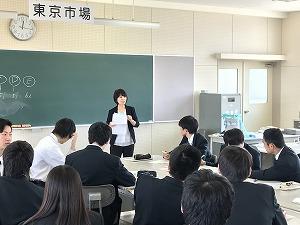 黒板の前で紙を手に話をしているスーツ姿の女性と、座って聞いている生徒たちの写真