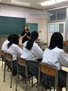 教室で、前方に立って笑顔で話す女性と、並んで座っている生徒たちの写真