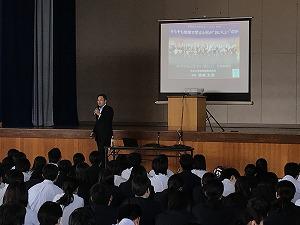 舞台の前で、スライド資料を背に話しているスーツ姿の男性と、座って聞いている生徒たちの写真