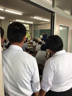黒板の前で授業をしている先生を、教室後方の入り口から見ている生徒たちの写真