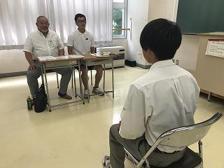 教室で、二人の男性面接官に向かって座り、模擬面接をしている男子生徒の写真