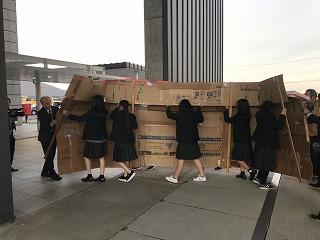 段ボールでできた壁を建物に運び込んでいる女子生徒たちと職員の人たちの写真