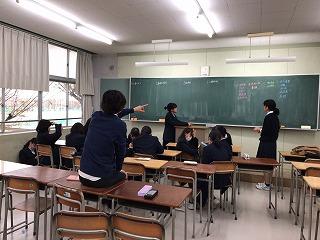 黒板の前に立っている二人の女子生徒に、机に座った人が後方から指示を与えている写真