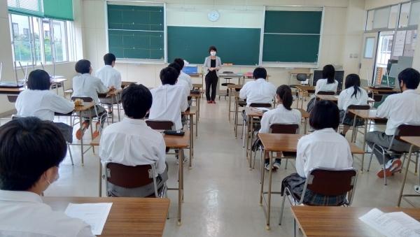 黒板の前で話をするスーツの女性と、着席して話を聞いている学生たちの写真