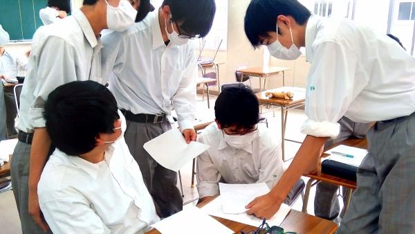 一つの机に5人の男子生徒が集まり、それぞれの紙を見せ合っている写真