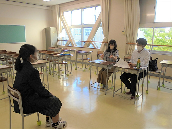 着席している二人の面接官が、少し離れたところに向かい合って座っている女子生徒に対して話をしてて、その面接の様子を2人の生徒が見学している写真。