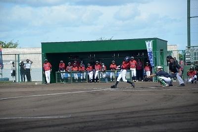 上半身が赤いユニフォームを着た選手たちがベンチにいるのを背景として一人の選手が打席でバットを振っている写真