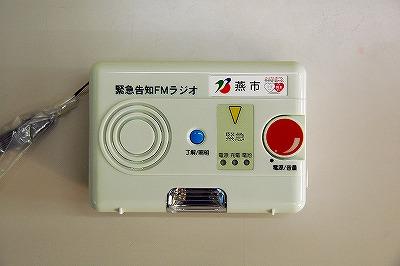 ストラップ付で、大きな赤い「電源/音量」ボタン、青い「了解/照明」ボタンがある「緊急告知FMラジオ」の写真