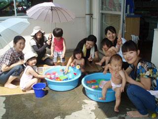 傘を差し屋外で水遊びをする幼児と保護たちの写真