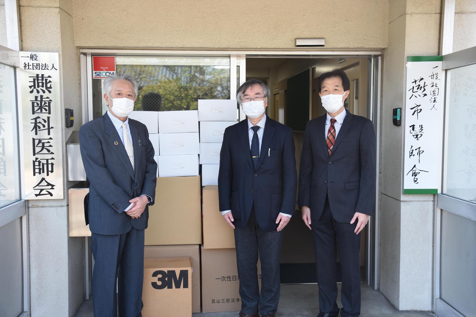 施設入口に積まれた段ボールとマスクを着用する役員たちの写真