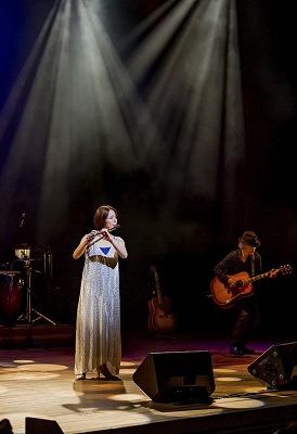 ステージの上で白いドレスを着てフルートを演奏する本宮氏の写真