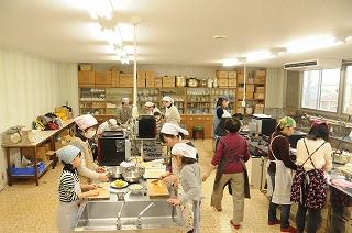 大きなステンレスの調理台が複数並ぶ部屋で調理をする、大人たちと子どもたちの写真
