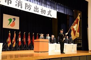 壇上で、大きな旗を掲げている制服制帽の男性と並べられた旗の写真
