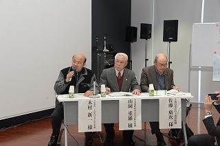 机を前に腰掛けて話をする3人の壮年の男性の写真