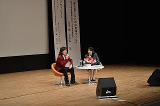 壇上の椅子に腰掛けて話すダイアモンド☆ユカイさんと女性准教授の写真
