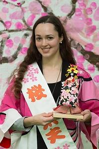人形を手に持ち、タスキを掛けて桜の柄の法被を着たロングヘアの外国人女性の写真