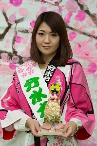 人形を手に持ち、タスキを掛けて桜の柄の法被を着たボブヘアの女性の写真