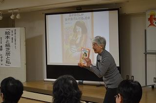 白いスクリーンの前で本を手にして話をするグレーの服を着た年配女性の写真