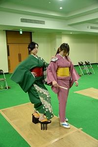 赤紫色の着物姿の女性の右肩に左手を乗せ高下駄で歩く緑色の着物姿の女性の写真