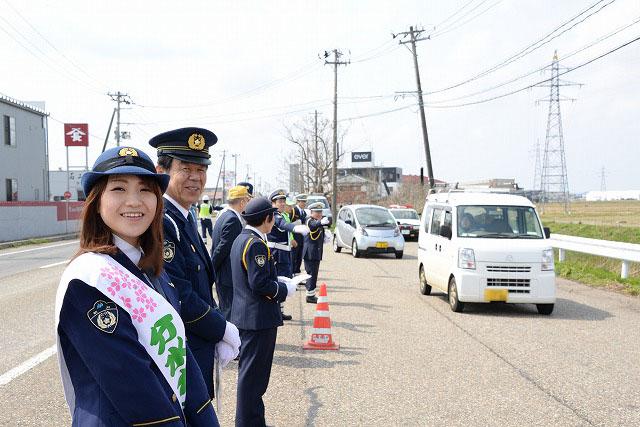 手前にいる笑顔の警察官の女性と笑顔の警察官の男性や、その奥に連なる警察官たちが道路を走る車のドライバーたちに呼びかけを行っている写真