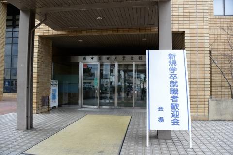 「新規学卒就職者歓迎会会場」と書かれたホワイトボードが入り口に立てかけられた吉田産業会館入口の写真
