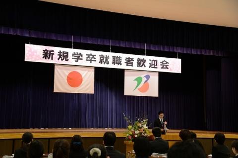 舞台に掲げられている「新規学卒就職者歓迎会」という文字が書かれた旗と、日の丸の国旗と燕市の市町村旗を写した写真
