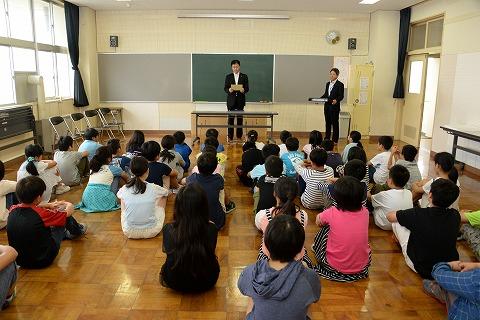 教室内で話すスーツ姿の男性と、床に座る子供たちの写真