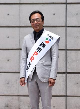 新潟県燕市PR大使のたすきをしている、メガネを掛けた笑顔のスーツ姿の男性の写真