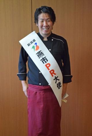 新潟県燕市PR大使のたすきをしているネイビーのシャツを着た笑顔の男性の写真
