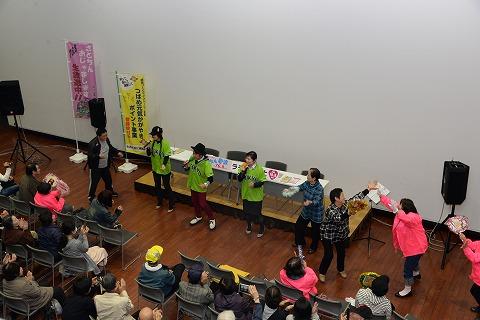着席して手を叩く観客の前で立ち上がりマイクを持ちながら体を動かす黄緑色のユニフォームを着た男女3人とピンク色のジャンパーを羽織った女性と向き合いながら両手を広げ合う高齢女性の写真
