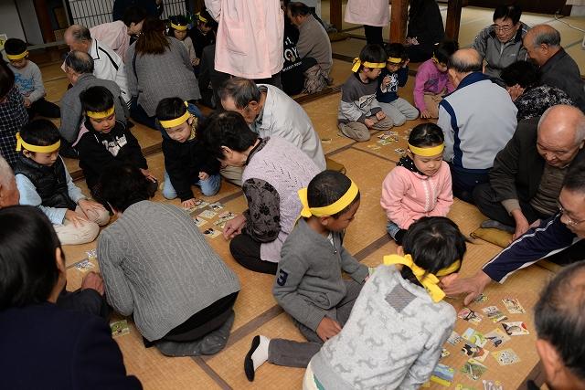 各々チームに分かれ畳の上に散らばった札を見つめたり札に手を伸ばしている黄色いはちまきをした子供たちと高齢男女たちの写真