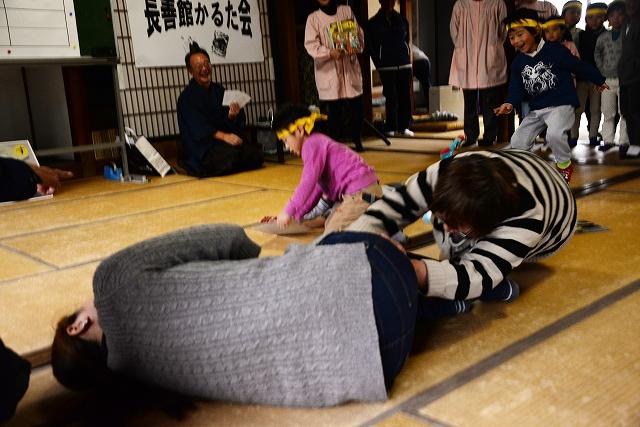 畳の上で倒れこむ2人の女性とその様子を見て札を手に口を開けて笑っている袴姿の男性の写真