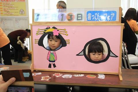 長机に立てかけられた和服を着たキャラクターとマスコットキャラクターの顔がくりぬかれたピンク色のボードの2つの穴に顔を突っ込む女の子の写真
