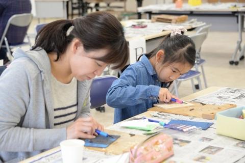 彫刻刀を使って絵が描かれた板を削るグレーのパーカーを着た女性と同じく板を削る青いシャツを着た女の子の写真