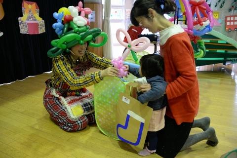 風船の帽子をかぶった人が、母親に支えられた子どもにピンクのバルーンを手渡している写真