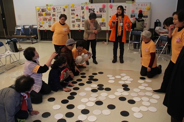 床に散らばったたくさんの白と黒2色の丸い紙を前にしゃがみ込む参加者たちと両手でジェスチャーをするオレンジ色の法被を羽織った女性の写真