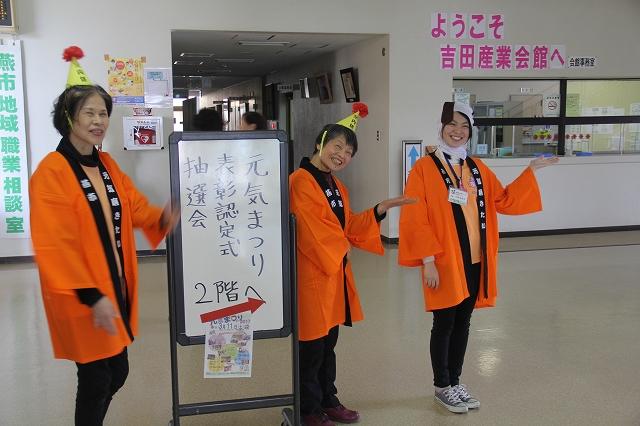 元気まつり表彰認定式抽選会と書かれた看板のそばで左手のひらを反し笑顔を浮かべるオレンジ色の法被を着た3人の女性の写真