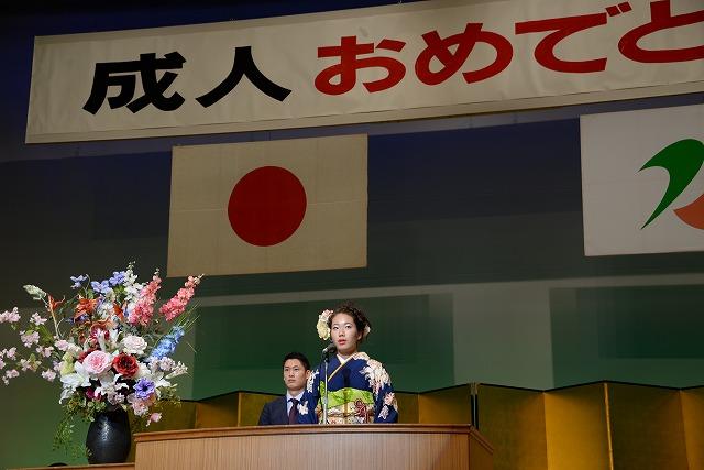 吊るされた成人おめでとうと書かれた横断幕と日本国旗と燕市のマークの下で壇上のマイクの前に立つ振り袖姿の女性と女性の後ろに立つスーツ姿の男性の写真