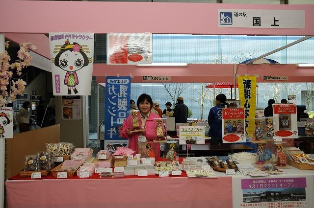テーブルにぎっしりと並べられた様々な商品がある道の駅国上と書かれたブース内でこちらを向きピンクの服を羽織った女性が販売されている人形を両手に持っている写真
