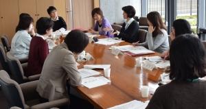 9人の女性が資料の置いてある四角い机を囲みながら会議をしている写真