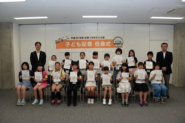任命書を手に持って記念撮影をする18人の子ども記者と後方両側に立つ市長とスーツの男性の写真
