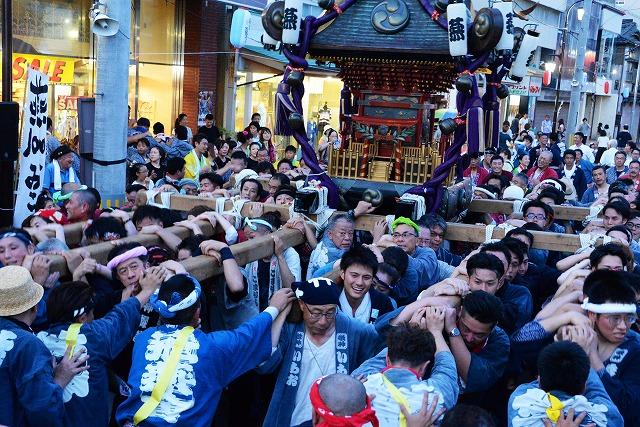商店街を征く、主神輿を担ぐ大勢の人々と参列客で賑わう様子を写した写真