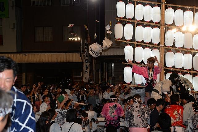 竿燈が取り付けられた神輿に乗っている女性が大入り袋の様な物を参列者にばら撒いる写真