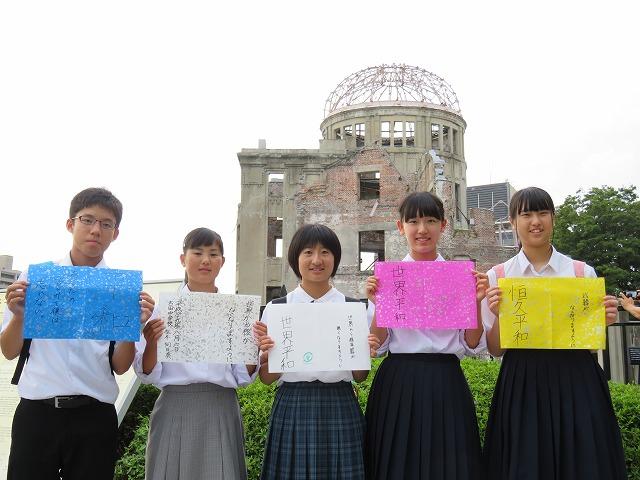 原爆ドームを背に「平和への決意」を記したカードを見せる5人の中学生の写真