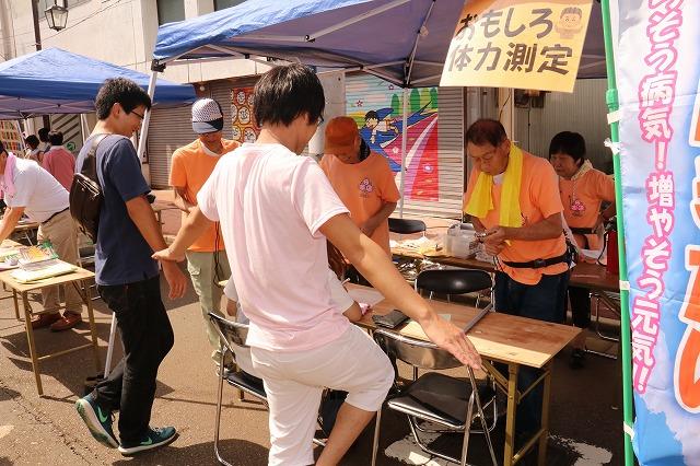 オレンジのTシャツを着た4人のスタッフと片足を上げてバランス測定をしている男性の写真