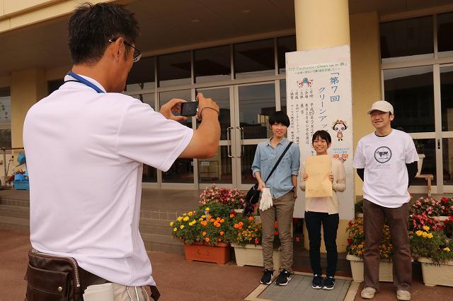 第7回クリーンアップ選手権大会の概要が書かれた紙の前で、カメラを持って撮影している男性とカメラを向けられて笑顔の男性と2人の女性の写真